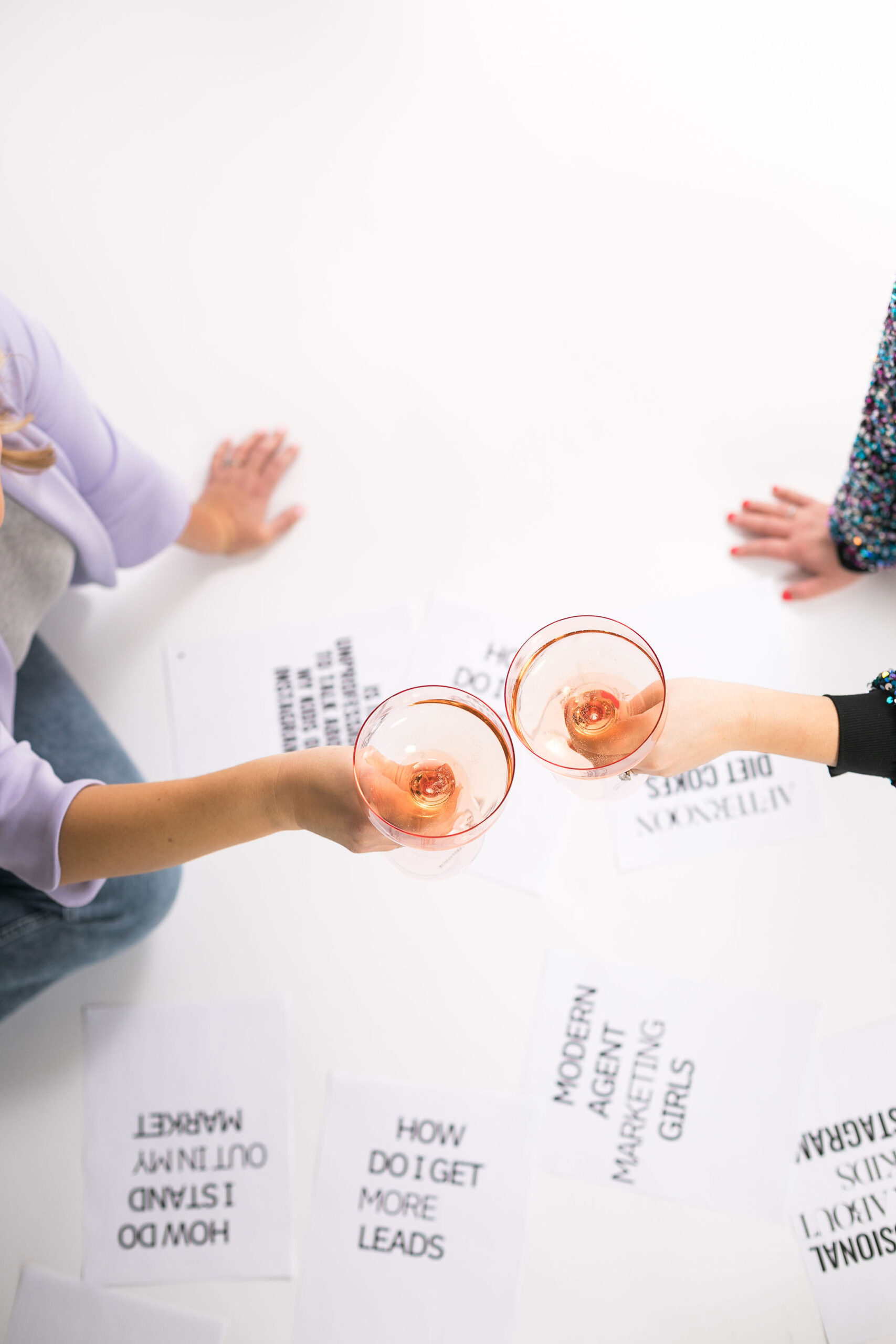 Chelsea and Kayla sitting on the floor with papers showing questions and holding two wine glasses