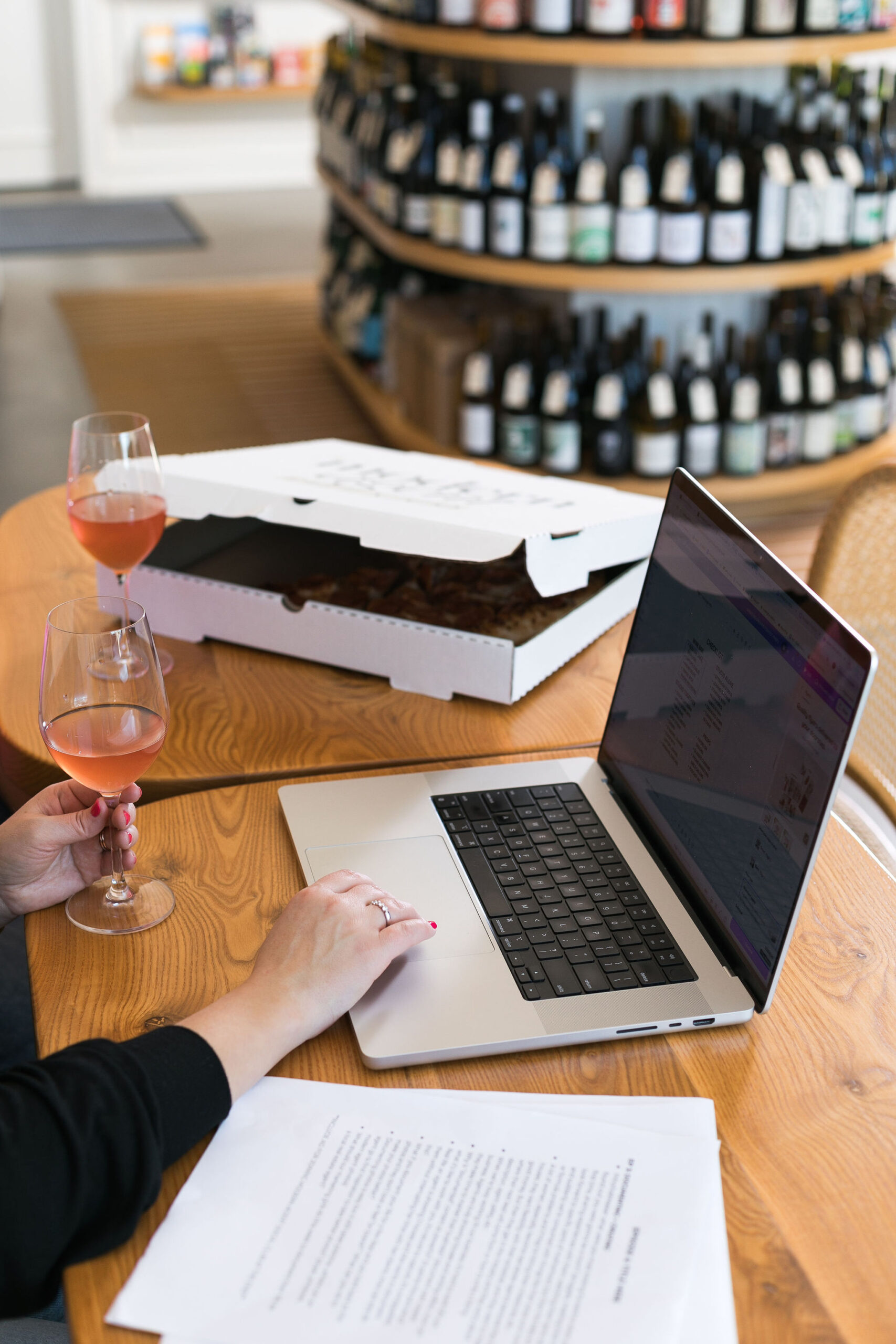 Hands typing on a laptop with a pizza box and wine glass nearby on a table in a wine shop
