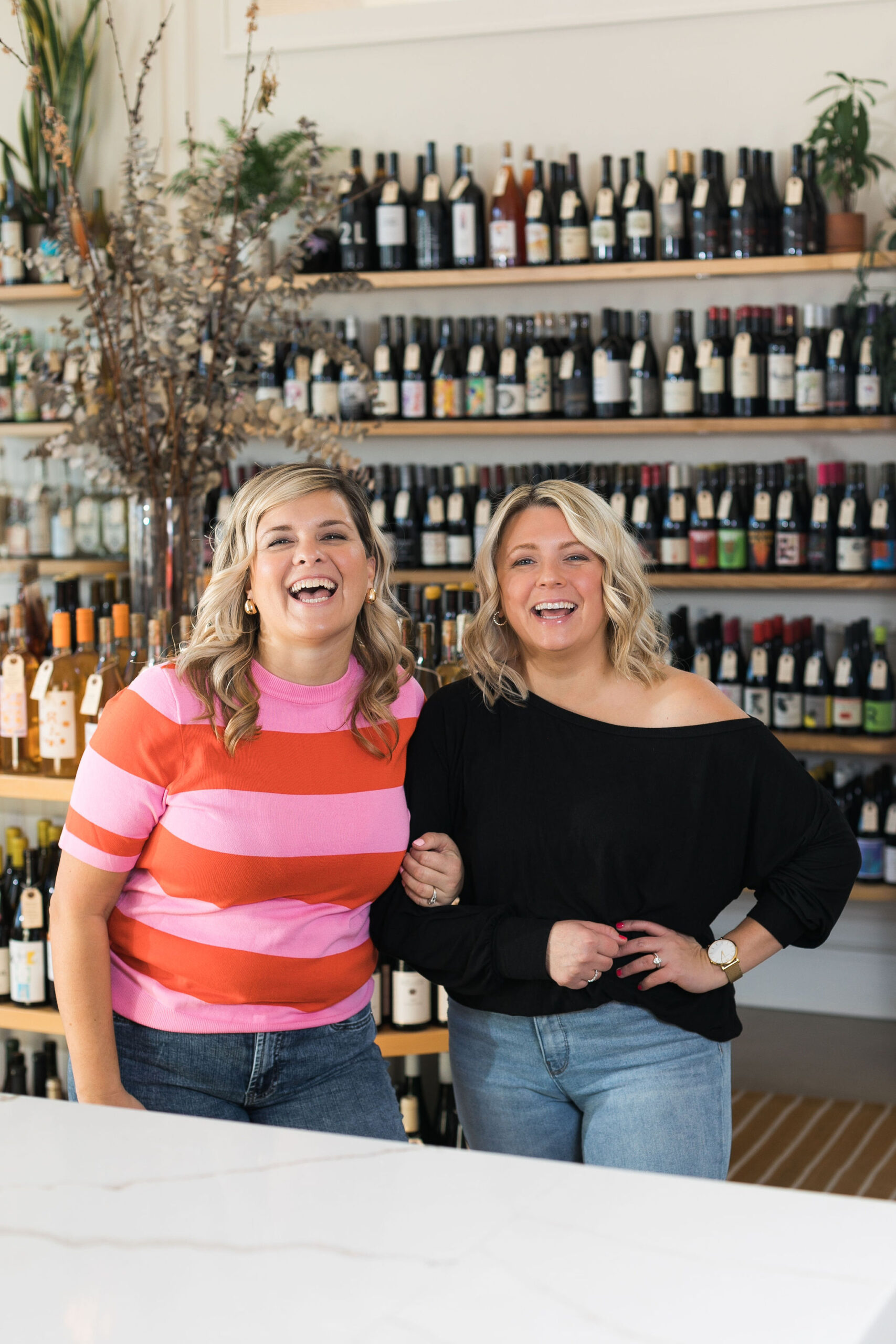 Chelsea and Kayla, arms linked, laughing, and standing in front of shelves of wine bottles