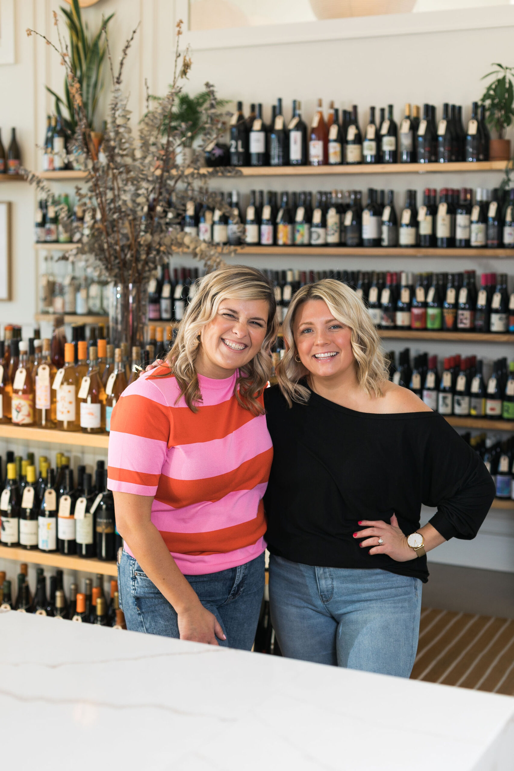 Chelsea and Kayla posing and smiling in front of wine shelves in a shop