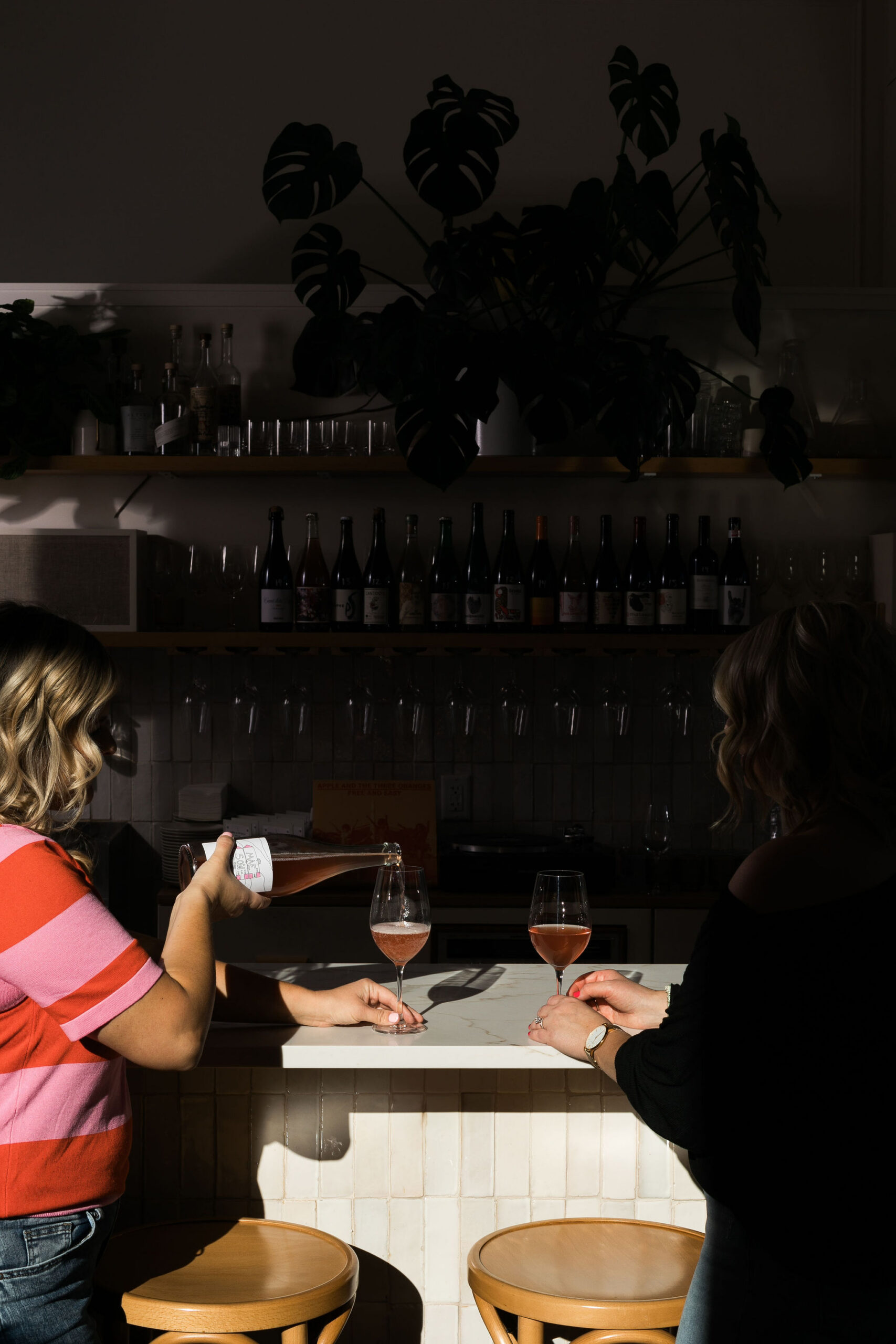 Chelsea and Kayla sitting at a wine bar, Kayla pouring wine into her glass
