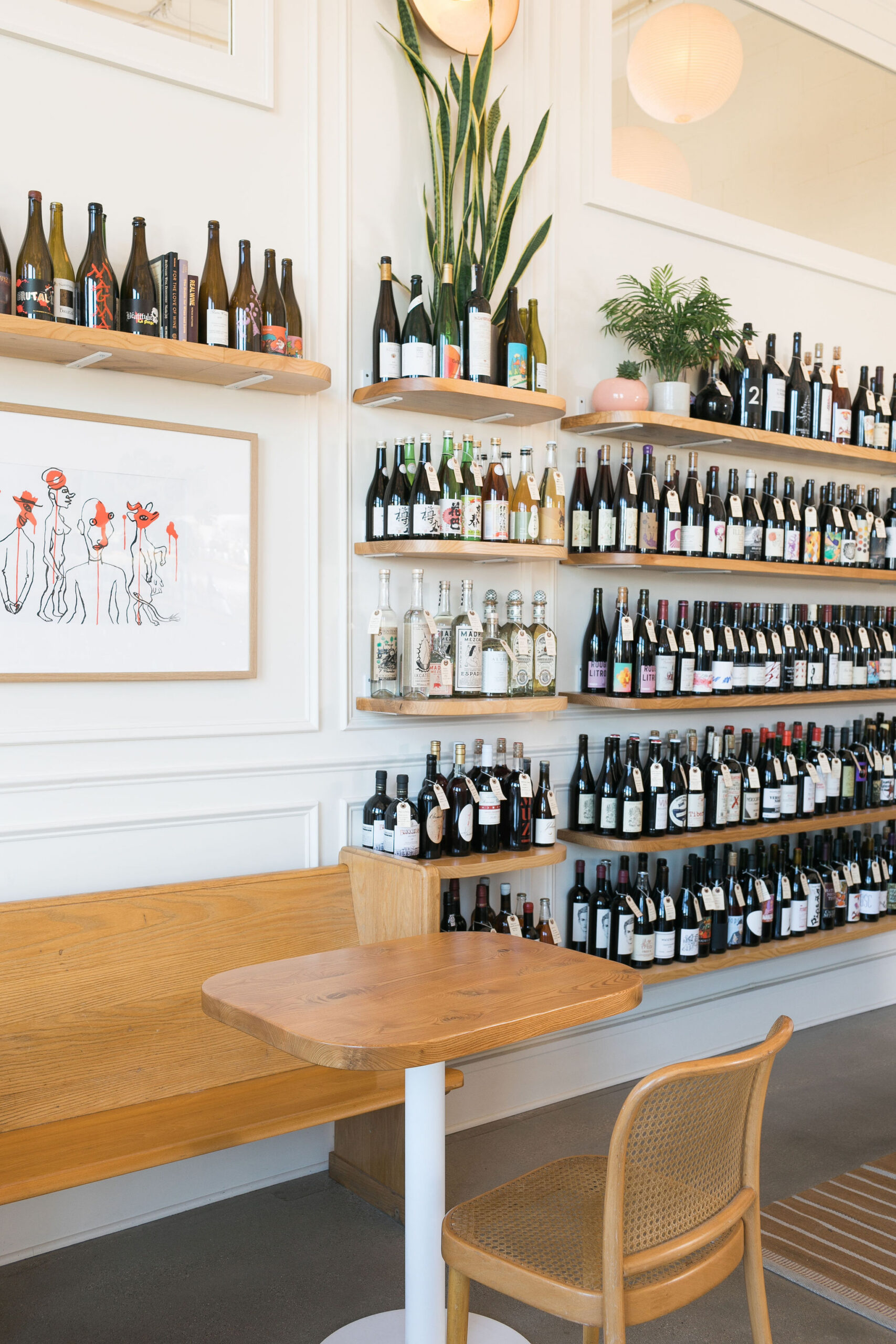 Cozy wine shop interior with wooden shelves full of diverse wine bottles, potted plants, framed artwork, and a wooden table with bench.